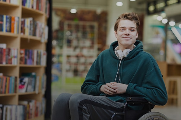 Νεαρός σε αναπηρικό αμαξίδιο χαμογελά με τα ακουστικά του περασμένα γύρω από το λαιμό, καθώς ετοιμάζεται να μοιραστεί τις ιδέες του για βελτίωση της εξυπηρέτησης των ατόμων με αναπηρία. Εικονίδιο προσβασιμότητας.